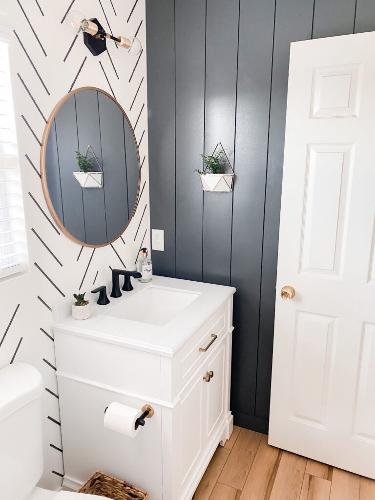 Kids Bathroom by popular San Diego interior design blog, Domestic Blonde: image of a remodeled kids bathroom with a white and grey stripe accent wall, modern lighting, round mirror, white vanity, light wood flooring, and abstract shape shower curtain. 