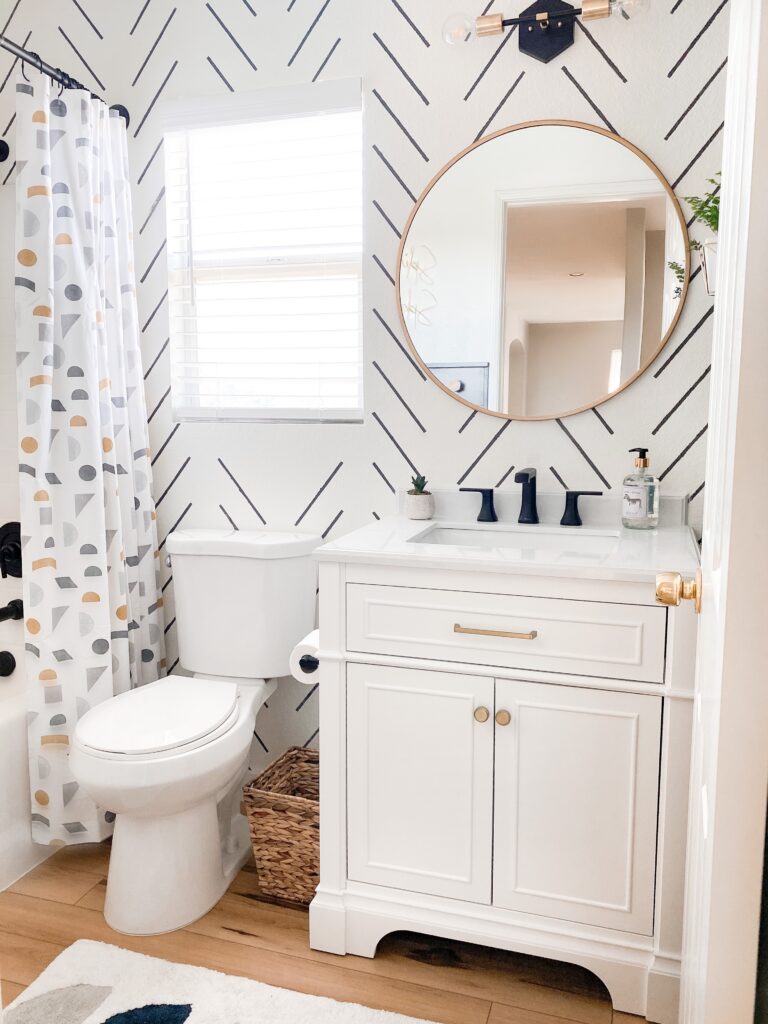 Kids Bathroom by popular San Diego interior design blog, Domestic Blonde: image of a remodeled kids bathroom with a white and grey stripe accent wall, modern lighting, round mirror, white vanity, light wood flooring, and abstract shape shower curtain. 