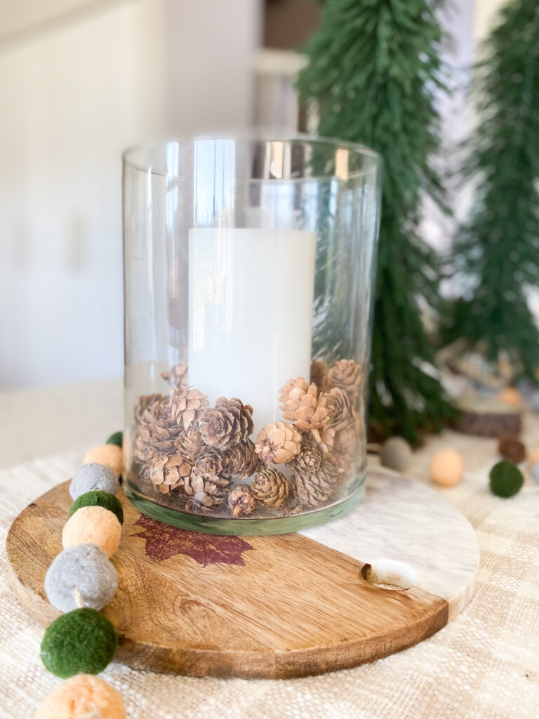 Joann Holiday Decor by popular San Diego life and style blog, Domestic Blonde: image of a wood and marble cutting board, pom pom garland, and glass jar filled with a pillar candle and mini pinecones.